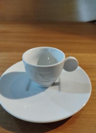 На картинке - белая фарфоровая чашка для кофе с блюдцем, относящаяся к бренду nespresso.