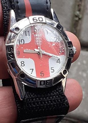 Наручные часы с циферблатом, на котором изображен флаг англии. у часов черный тканевый ремешок и серебристый корпус.