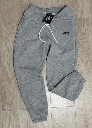 Це сірі спортивні штани stussy з вишитим логотипом бренду. вони виготовлені з бавовняного флісу та мають вільний крій, що забезпечує комфорт