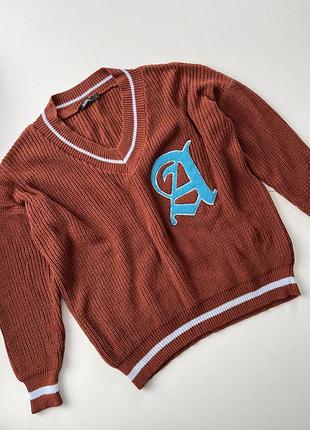 Коричневий в'язаний светр з v-подібним вирізом, білими смужками та блакитною нашивкою у формі літери "а"