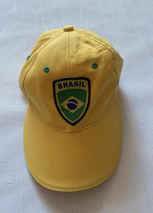 Це яскраво-жовта бейсболка з вишитим щитом і написом "brasil", схожа на товари для фанатів футбольної збірної бразилії.