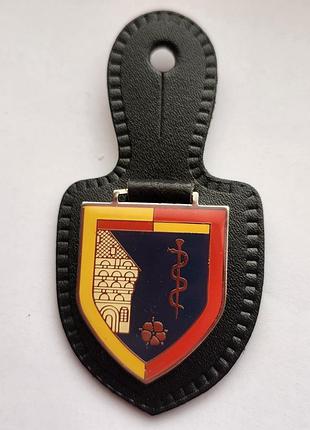 Колекційний знак, ймовірно, пов'язаний з медичною службою або відповідною організацією.  предмет: емальований значок у формі щита на шкіряній основі.