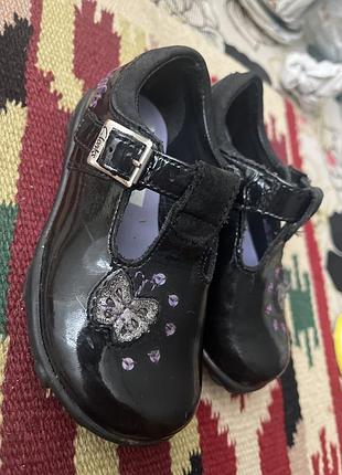 Дитячі лаковані туфлі бренду clarks у чорному кольорі, що застібаються на ремінець.