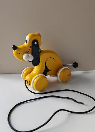Деревянная каталка brio в виде персонажа disney — плуто.игрушка имеет желтый корпус, белые колеса и веревочку для катания. деревянна каталка