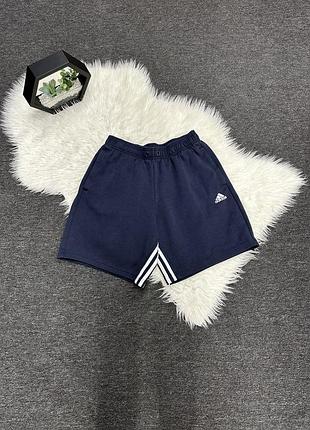 Темно-сині спортивні шорти adidas з білим логотипом на лівому стегні та фірмовими смужками на розрізах знизу.