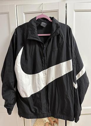 Чорна вітрівка nike із великим білим логотипом swoosh на грудях та рукаві виконана у стилі оверсайз із нейонового матеріалу