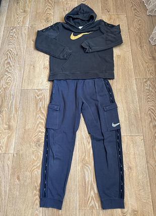 Спортивний костюм nike із серії repeat, що складається з темного худі з золотистим логотипом swoosh та синіх штанів-карго з фірмовими лампасами-лого.