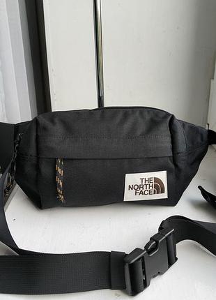 Черная поясная сумка the north face lumbar pack с винтажным тканевым логотипом и оранжево-черным шнуром на переднем кармане.