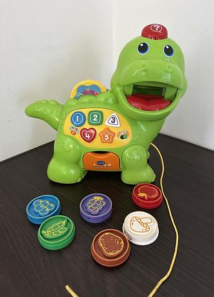 Розвиваючий динозавр діно vtech chomp and count dino зеленого кольору з різнокольоровими фішками-жетонами у вигляді їжі.