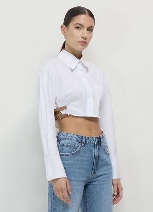 Укороченная белая рубашка бренда zara с длинными рукавами, накладным карманом и декоративным ремешком с пряжкой на боку.