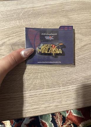 Вінтажний металевий значок-пін (lapel pin) із золотистим покриттям та червоними елементами, що є туристичним сувеніром із надписом «visit malaysia».