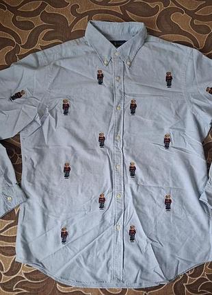 Это светло-голубая хлопковая рубашка oxford от polo ralph lauren, украшенная фирменным принтом-вышивкой с городскими мишками.