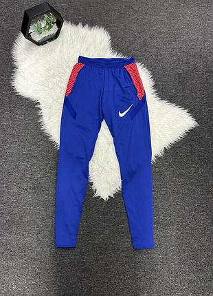 Сині спортивні штани nike dri-fit strike з червоними вставками на стегнах та білим логотипом бренду.