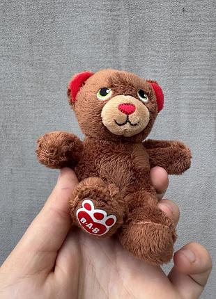 Маленька плюшева іграшка-ведмедик бренду build-a-bear із фірмовим логотипом «b-a-b» на стопці