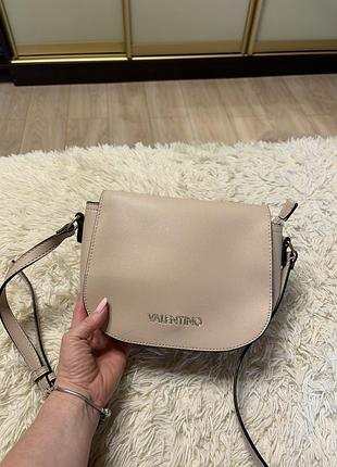 Бежевая сумка-кроссбоди от valentino с округлым клапаном и металлической надписью спереди.