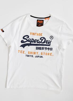 Біла, чоловіча футболка superdry з виразним, вінтажним принтом у фірмовій стилістиці бренду. на передній частині великий напис superdry у темно-синьо