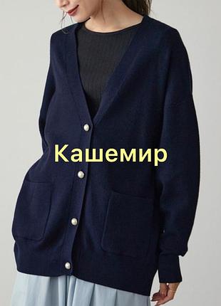 На зображенні представлений подовжений темно-синій кардиган вільного крою з v-подібним вирізом, передніми гудзиками та накладними кишенями.