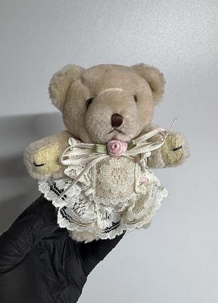 Вінтажний плюшевий ведмедик у світлому кремовому відтінку, одягнений у декоративну білу сукню або фартух із мережива та прикрашений маленькою рожевою