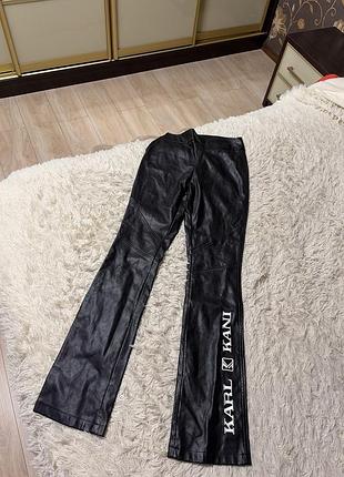 Чёрные брюки из экокожи с крупным белым логотипом karl kani на левой штанине.